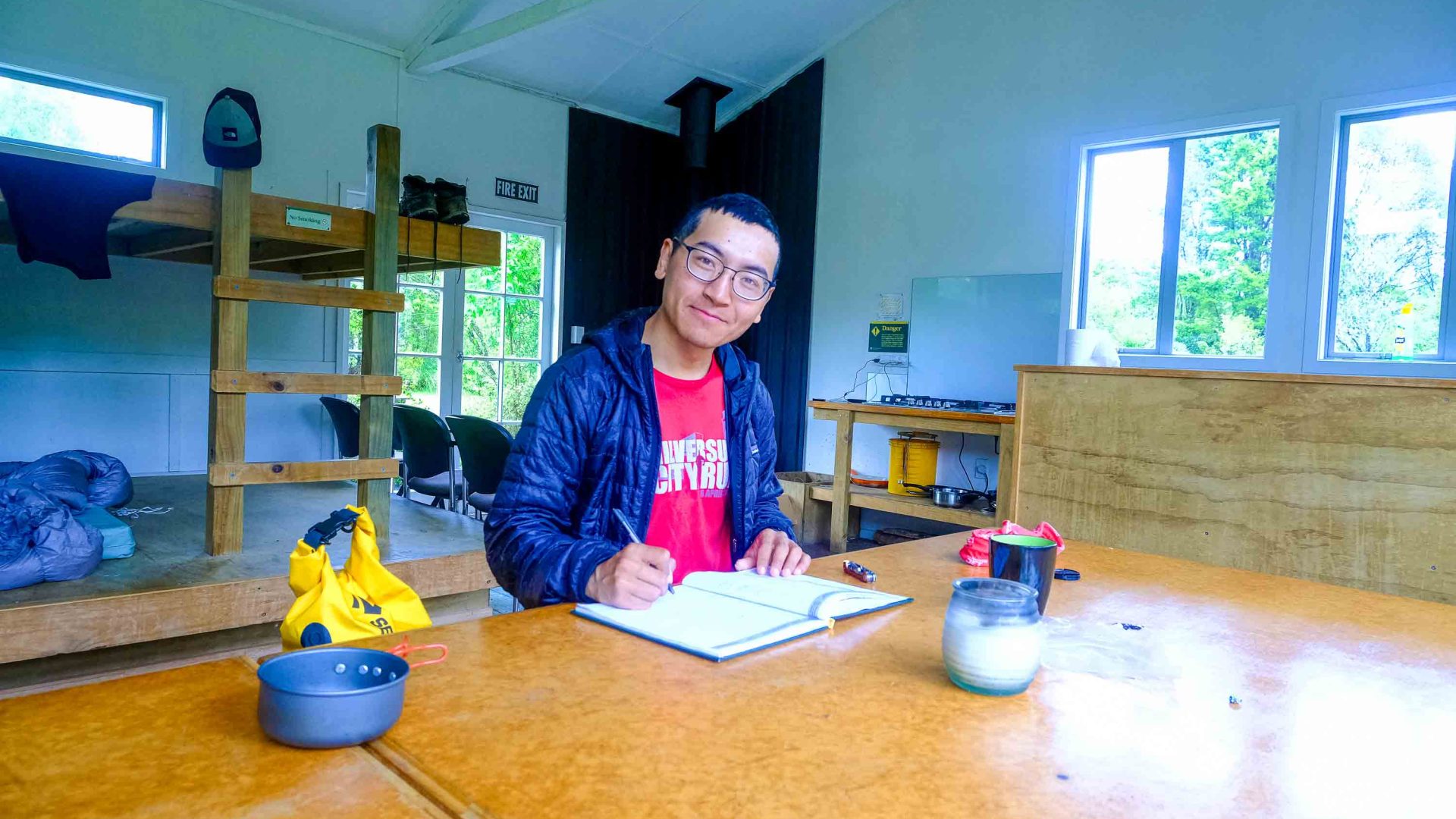 The writer adheres to the tradition of writing in the book which is at every hut, to record details from the trail. He sits inside a hut and looks at the camera with the book in front of him.